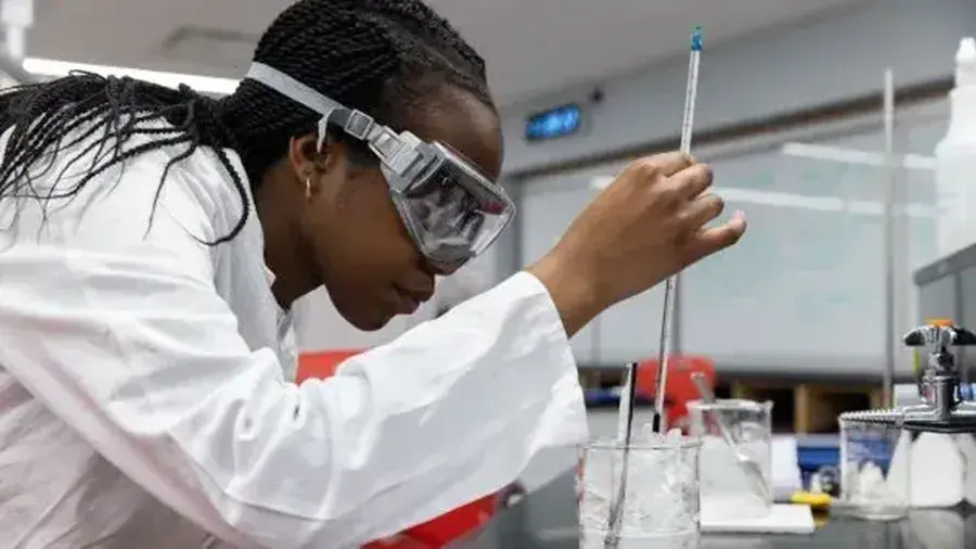 A person in a white lab coat works at a laboratory bench, holding a glass instrument over a beaker filled with liquid, with other lab equipment and a notebook nearby.
