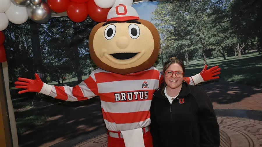 Elizabeth stands next to Brutus Buckeye with a campus back drop behind them.