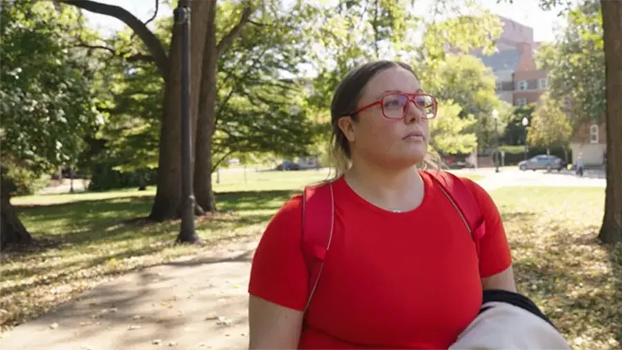 Elizabeth wearing a red shirt and backpack standing on a tree‑lined sidewalk in a park, holding a light‑colored jacket, with buildings and parked cars in the background.