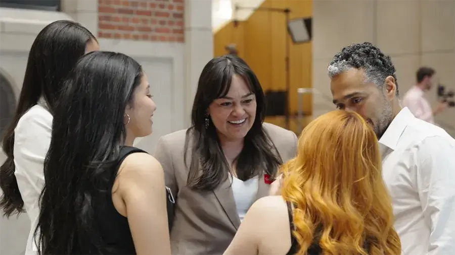 Five people stand in a small circle, facing inward as if talking. They are indoors in a large room with brick and light-colored walls, studio lights, and equipment visible in the background.