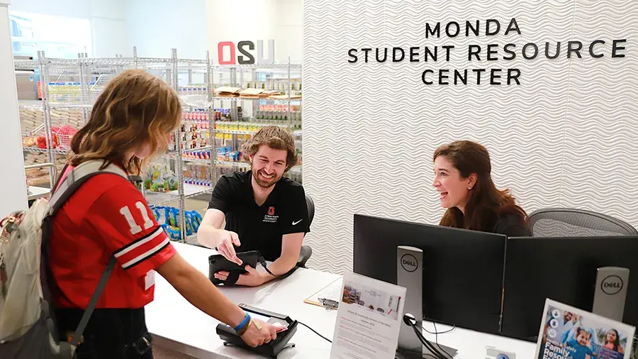 A person stands at a service counter at the Monda Student Resource Center, handing an item to staff seated behind computer monitors. Shelves of food and supplies are visible behind the desk, with “OSU” and “Monda Student Resource Center” signage on the wall.