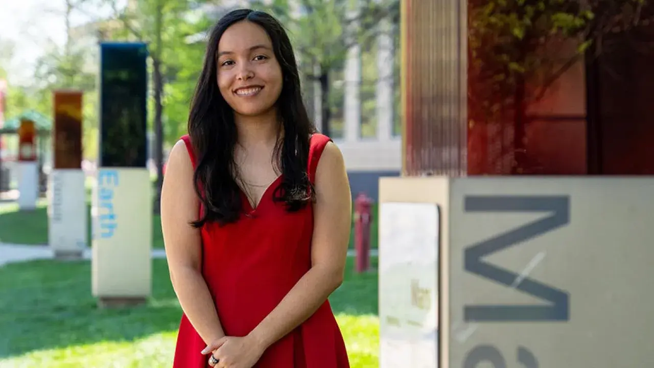 Hannah wearing a red sleeveless dress stands on grass outdoors, with hands clasped, in front of large rectangular art panels and trees.