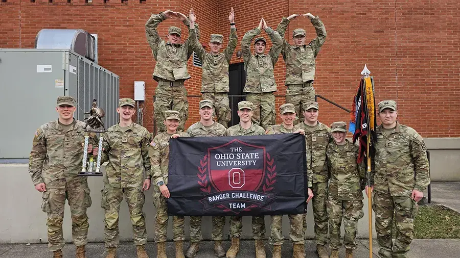 Ohio State University Ranger Challenge Team in military uniforms posing with a team flag and trophy outside a brick building.