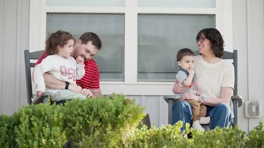Two adults seated on a front porch each hold a young child while sitting in rocking chairs. Shrubs fill the foreground, and large windows are visible behind them.