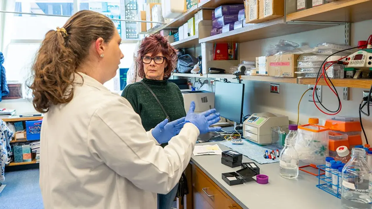 Person in a lab coat and gloves speaks with another person in a laboratory workspace filled with equipment and supplies.