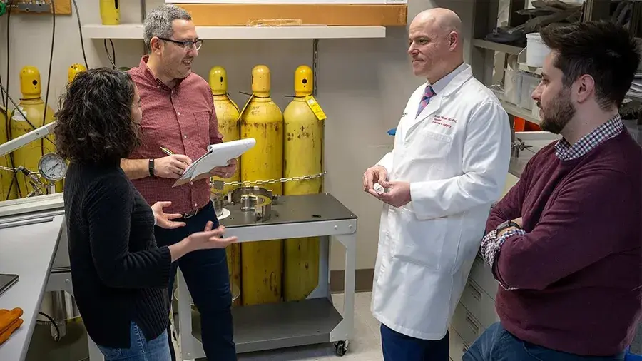 Four people standing in a laboratory, engaged in discussion near scientific equipment and yellow gas cylinders.