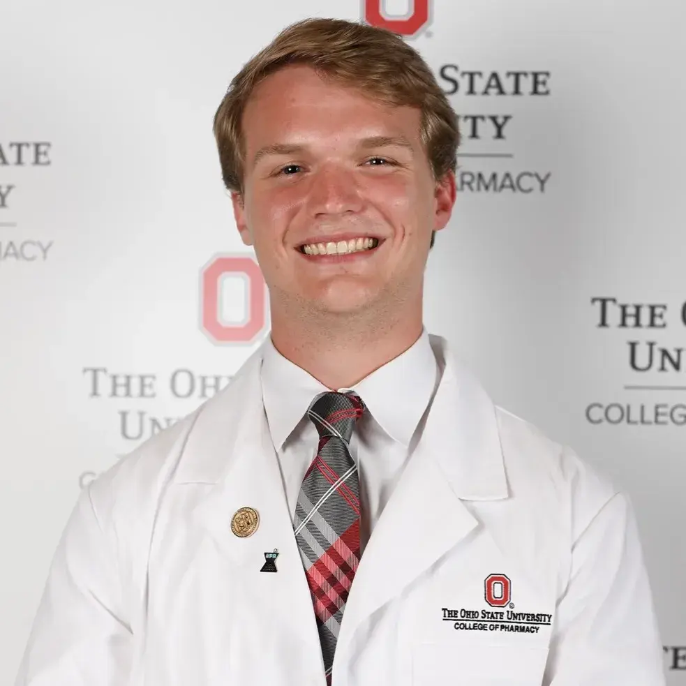 A white man with blonde hair wears an Ohio State white pharmacy lab coat and smiles