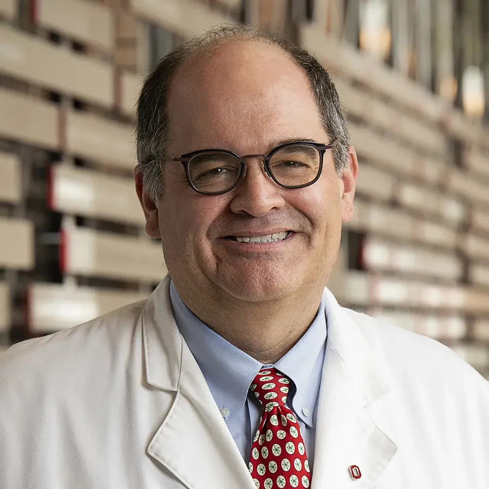 Andrew Thomas smiles big and wears glasses and a white physician lab coat