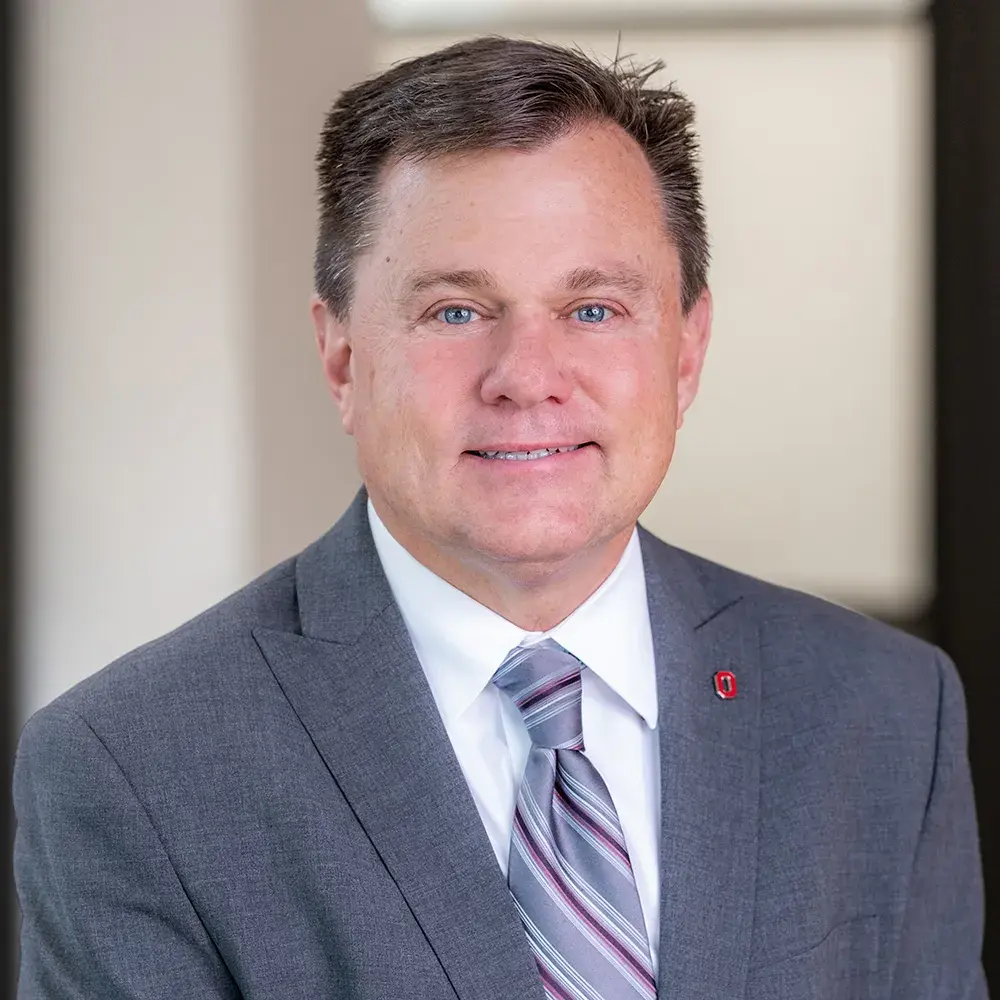 John Woods smiles and wears a gray suit with a scarlet Block O lapel pin