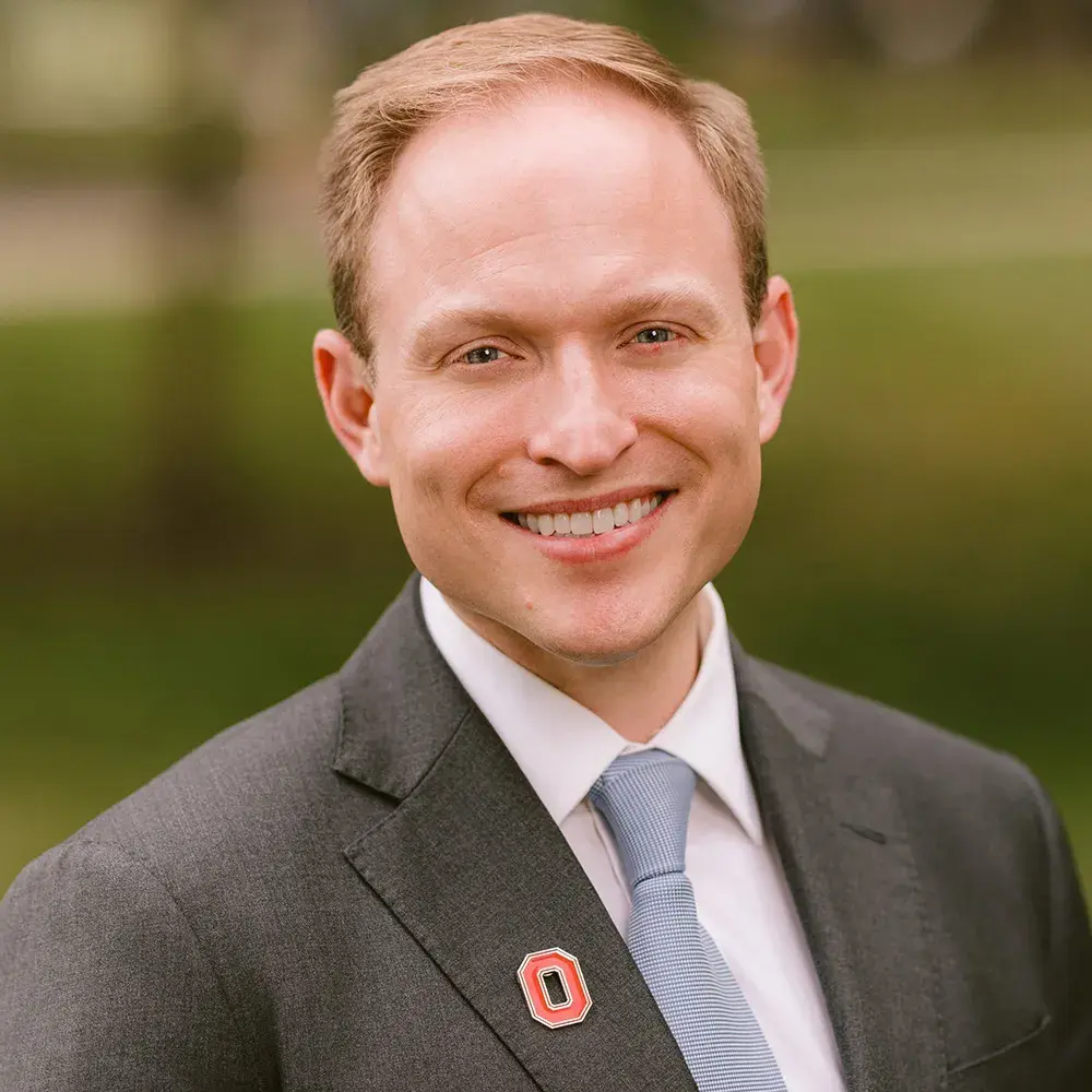 Kent Barnett stands outside and smiles big wearing a gray suit and Block O scarlet lapel pin