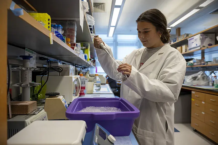 A woman wearing a white lab coat stands in a research lab doing an experiment