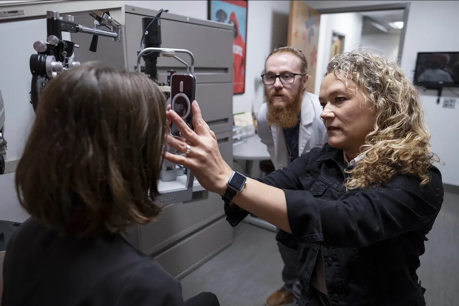 A woman performs an eye exam on a patient while a man looks on from behind