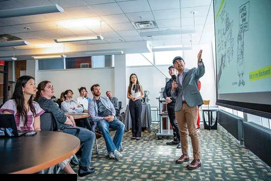 A group of students listen to a speaker who is presenting information on a screen