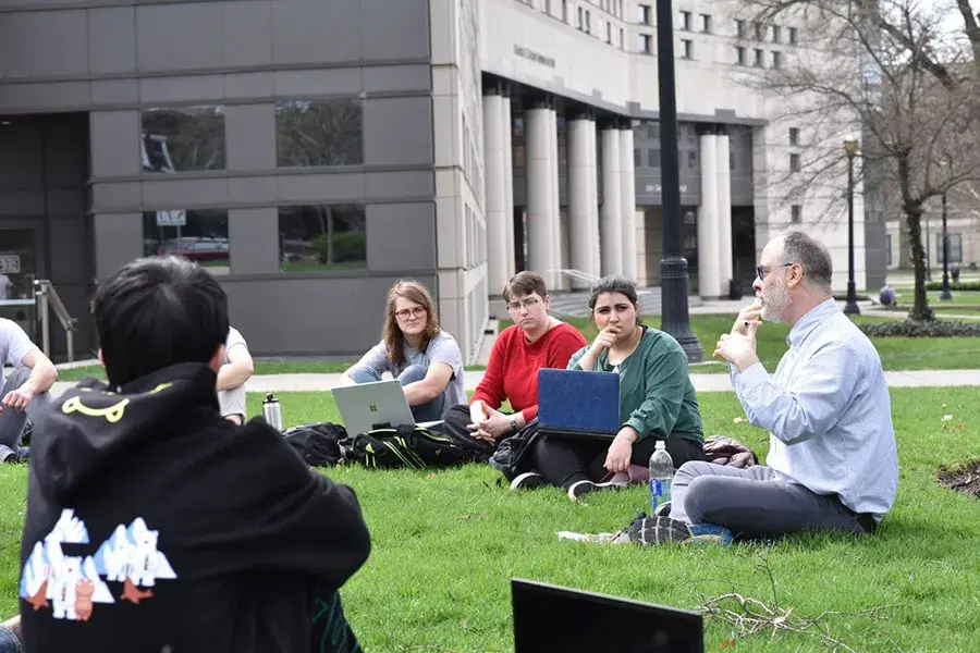 A group of students sit in a circle in the grass while listening to their professor speak