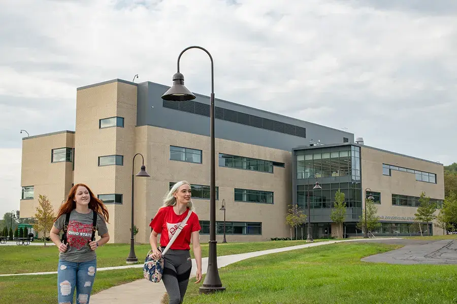 Two women smile as they walk on a sidewalk with a campus building behind them