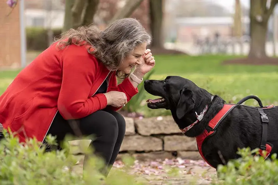 An older woman with long, gray hair wearing a red jacket sits on a stone wall and leans in toward a black dog smiling.