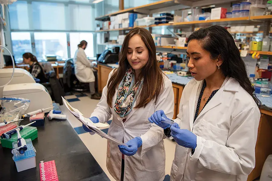 Women researchers stand in a lab wearing white lab coats and look at experiment papers