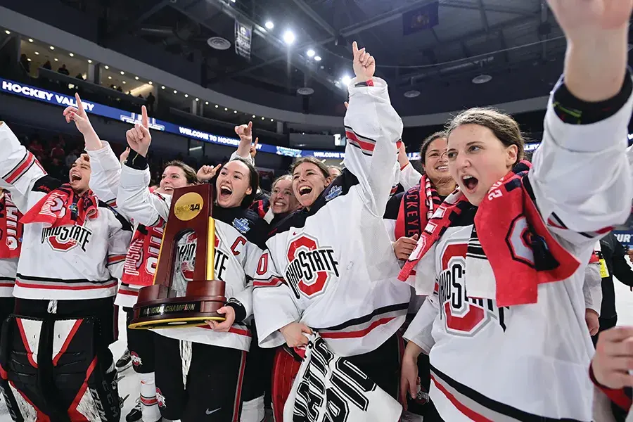 A group of Ohio State hockey players celebrate on the ice while holding a championship trophy, raising their arms in victory.