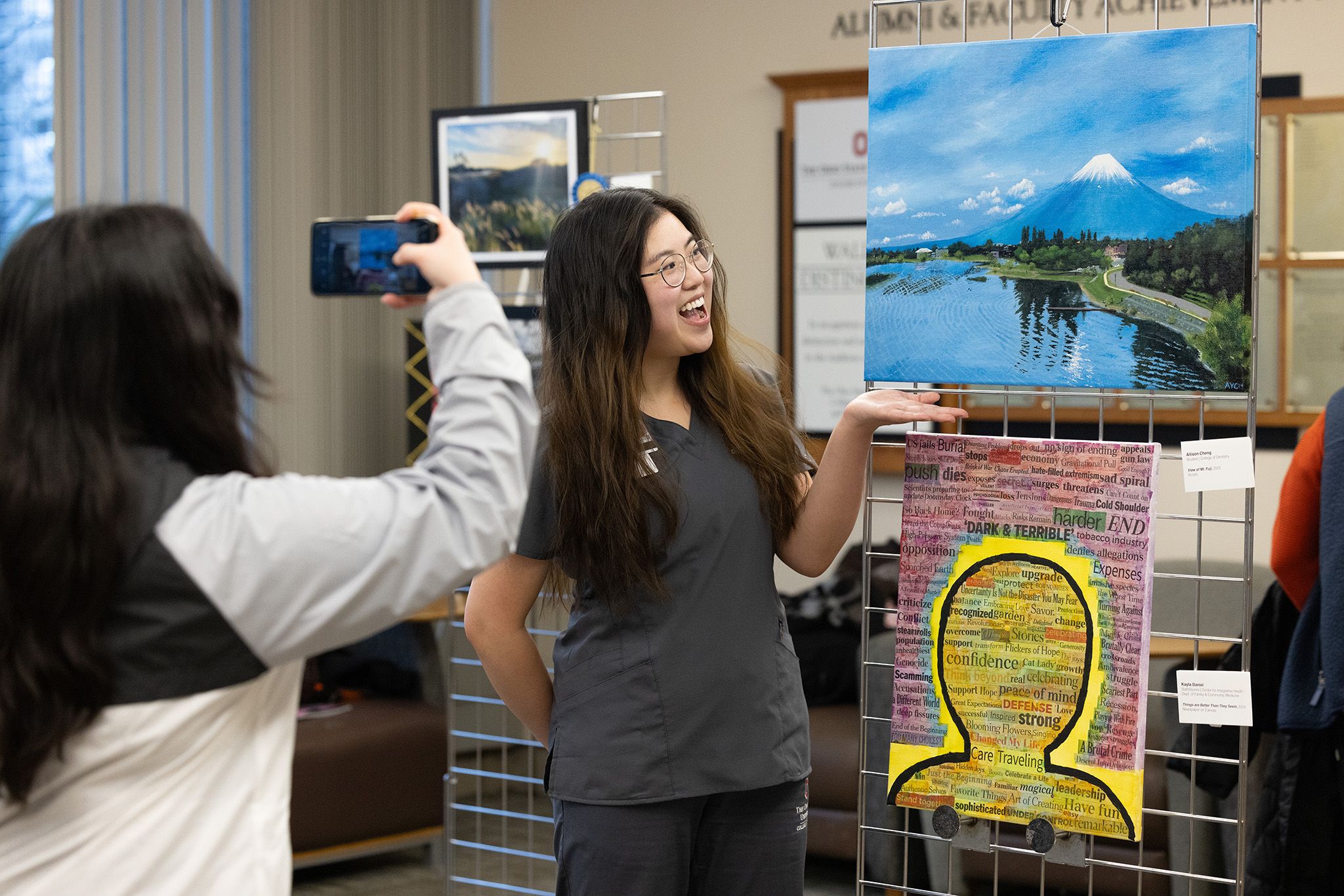 Beaming woman having her photo taken in front of her art, a painting of a mountain scene