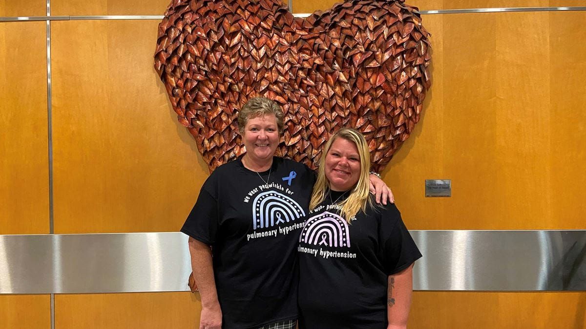 Two women stand in front of a large heart-shaped piece of artwork.