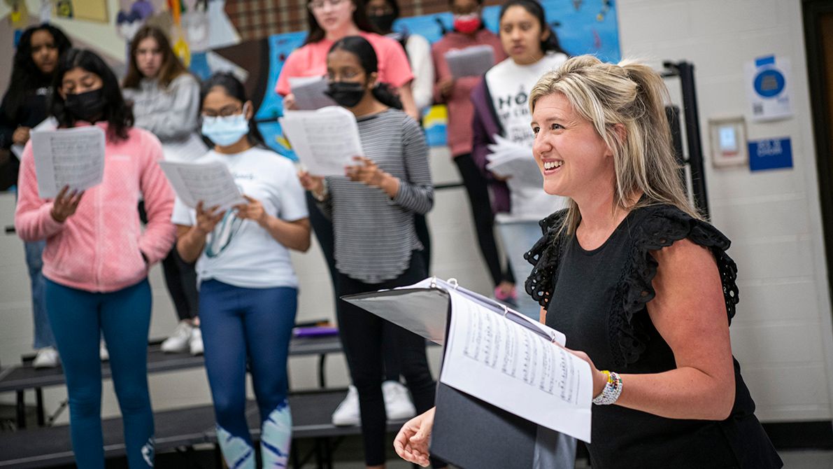 Teacher Katie Silcott smiles while holding sheet music as students stand in the background