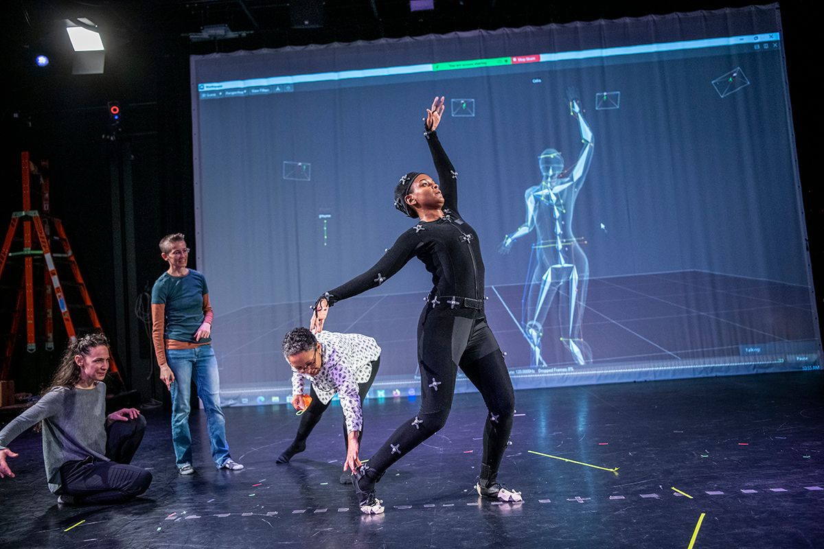 A dancer in a motion capture suit performs movements that are displayed on a digital screen behind her as three women faculty members look on.