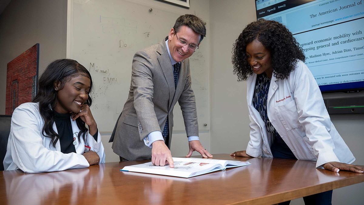 Two women in lab coats and a man in a suit in discussion at a table.