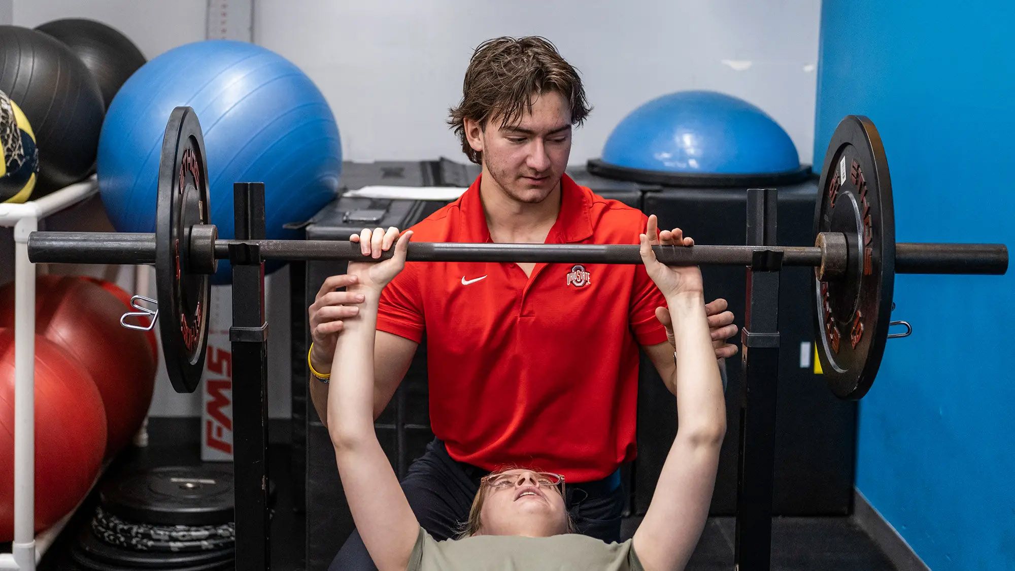 At a fitness gym, a young man with messy hair focuses on guiding a young woman getting ready to lift a weighted bar for a bench press. The man sits behind her, as she lies back on the bench and looks at the bar. He barely touches her wrists.  