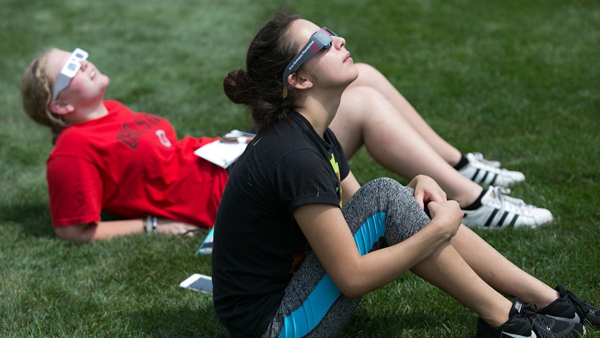 two people laying and sitting in grass look up with protective eyewear