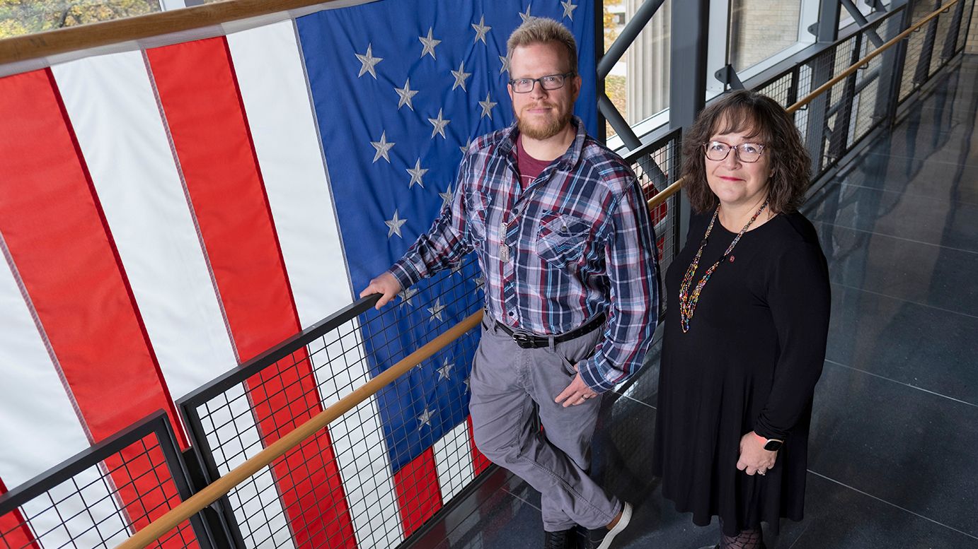 A man and a woman stand in front of a large American flag.