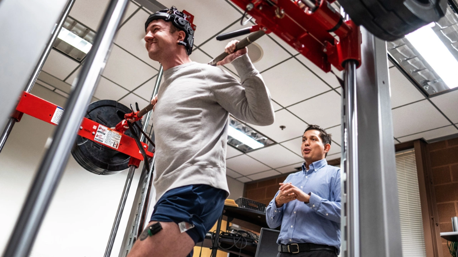 A man lifts weights with sensors attached, while another man observes.