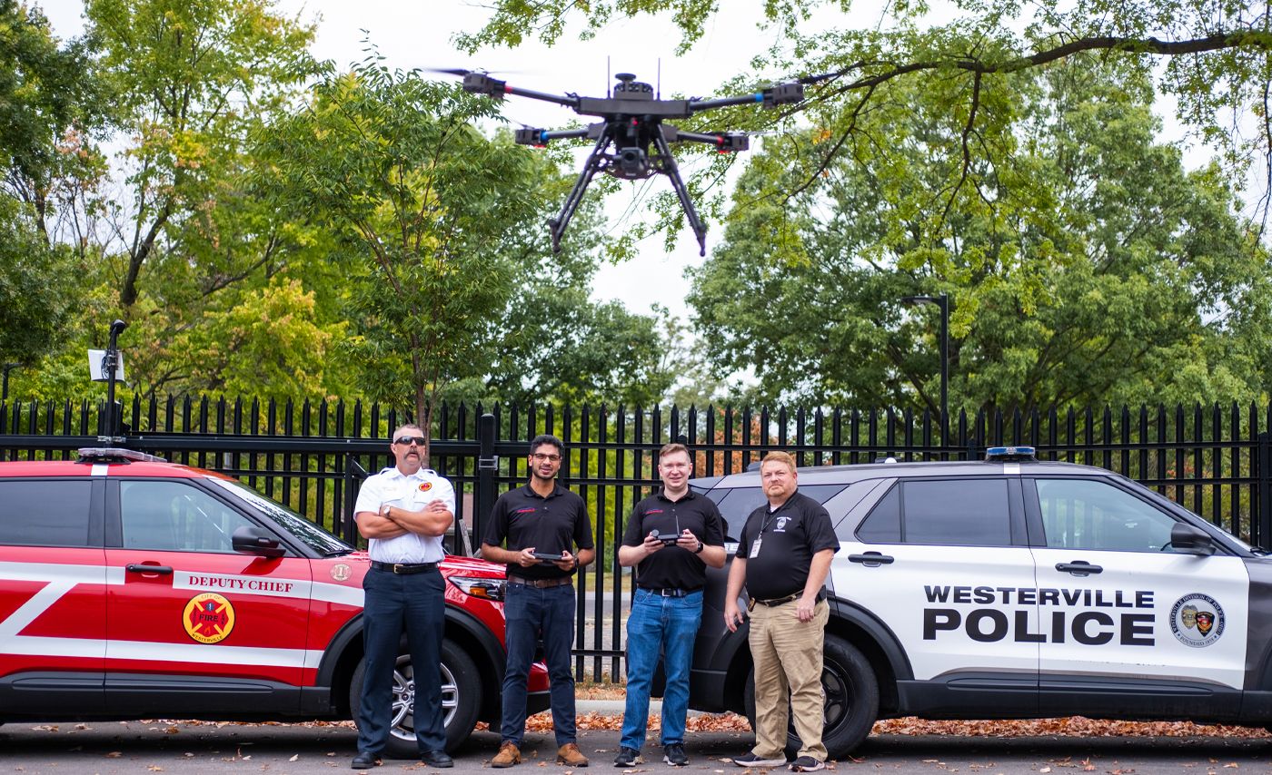 A drone flies in front of two students and two first responders.